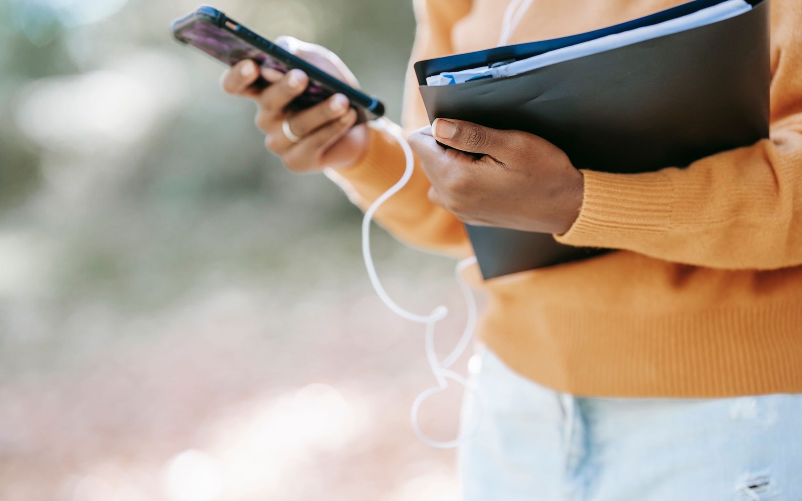 Hands checking a smartphone while holding a folder of paperwork outdoors