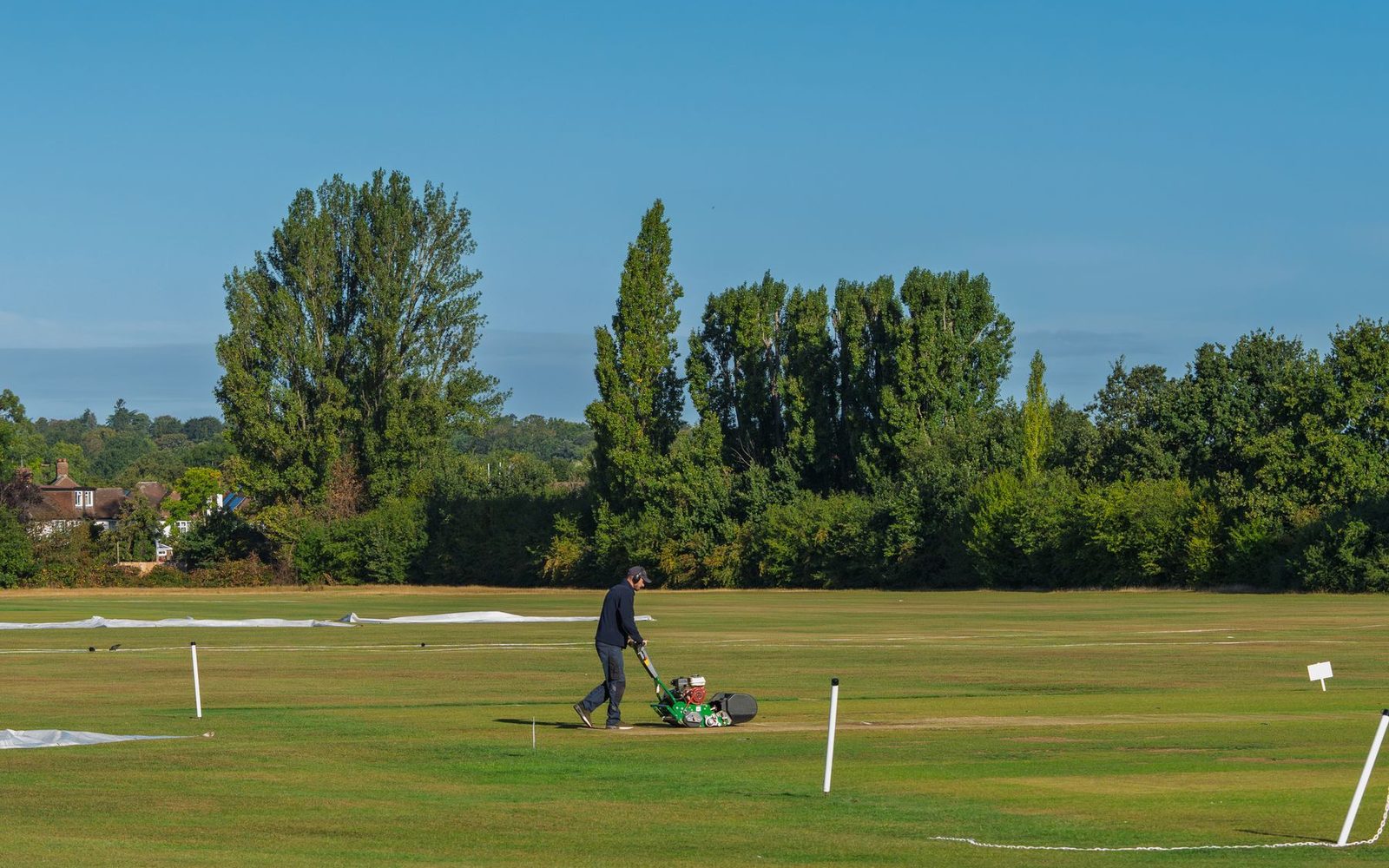 Groundskeeper pushing a mower across a sunlit lawn bordered by mature trees