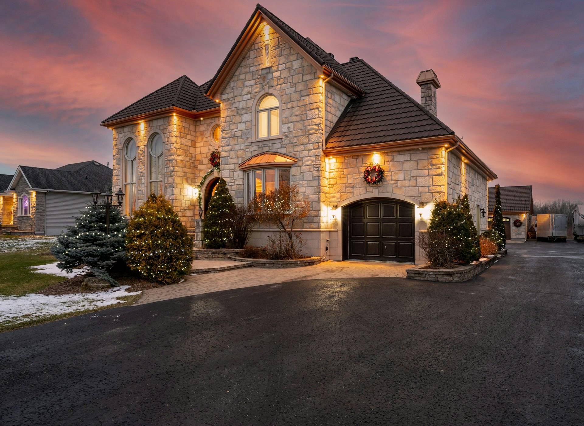 Stone house at dusk with warm lights glowing in the windows and evergreen shrubs along the front beds