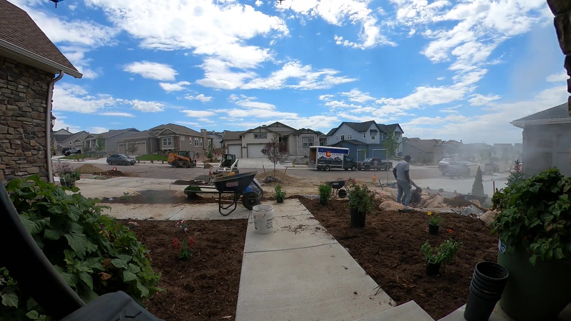 Landscaping crew planting new front-yard beds with fresh mulch, a wheelbarrow and a skid steer on a sunny day